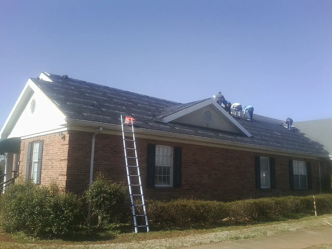 Crew tearing shingles off a brick ranch roof with a ladder set — Metal Roof Repair work by our crew in Campbell Hall