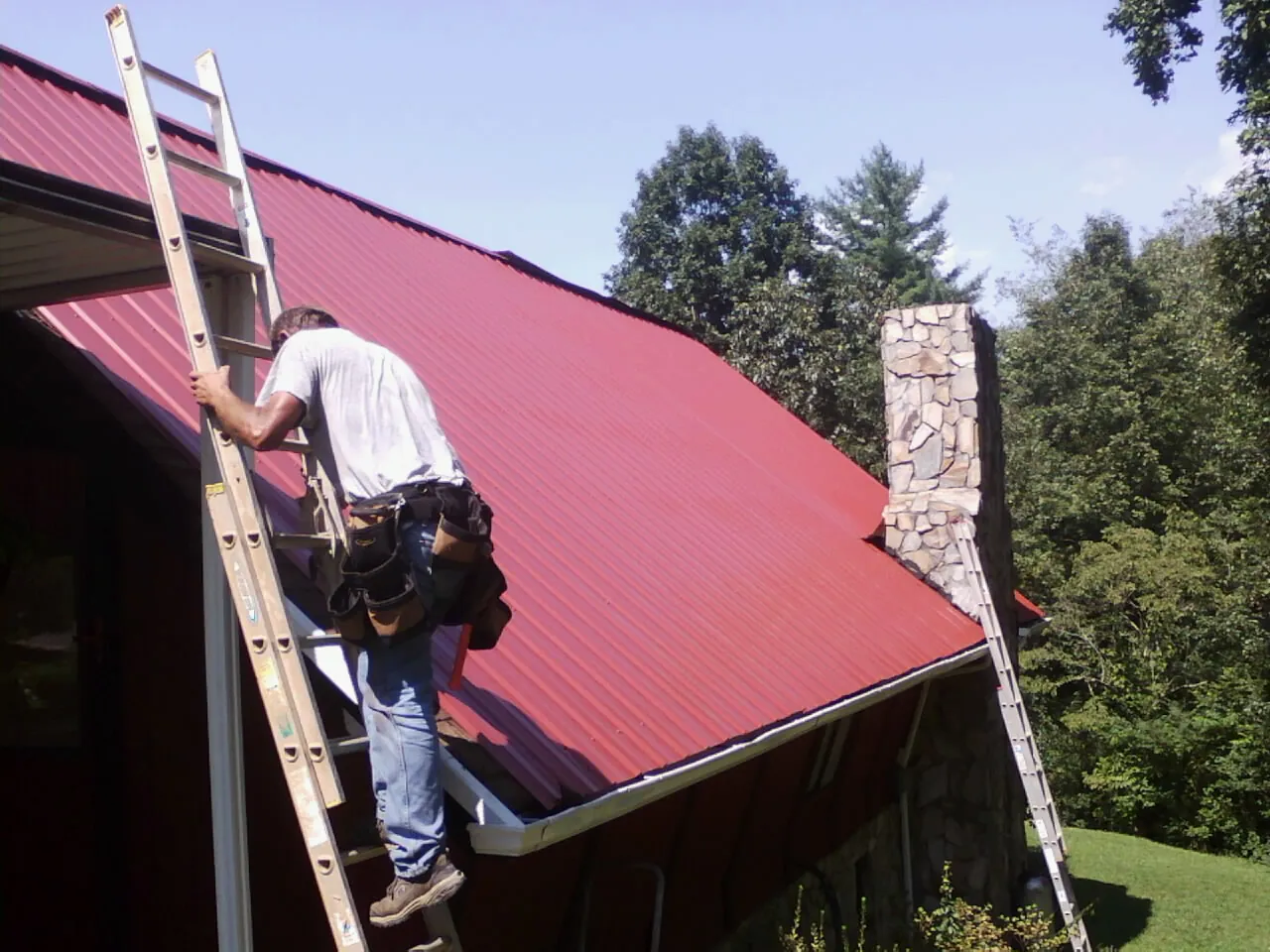 Roofer on a ladder beside a red metal roof and stone chimney — Soffit & Fascia Repair by Roofing Brothers in Campbell Hall